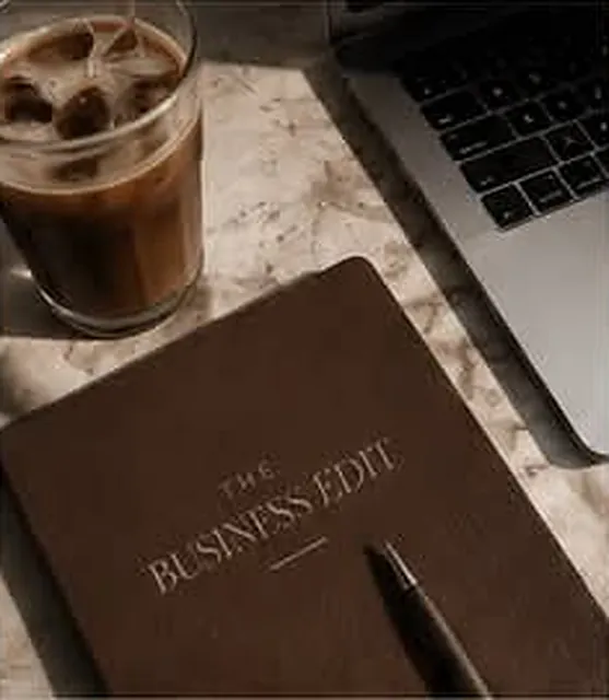 Espresso-brown notebook with The Business Edit logo beside an iced coffee and laptop